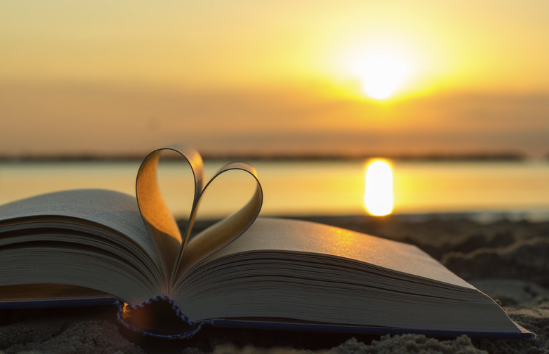 an image of a book open on a beach with two of the pages folded into a heart with a sunset in the background